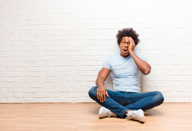 Young man looking sleepy, bored and yawning, with a headache and one hand covering half the face sitting on the floor at home