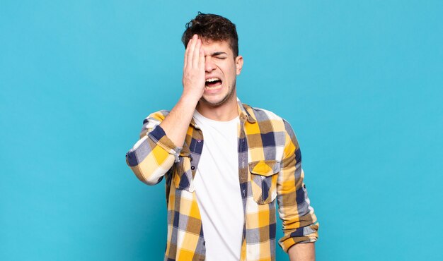 Young man looking sleepy, bored and yawning, with a headache and one hand covering half the face