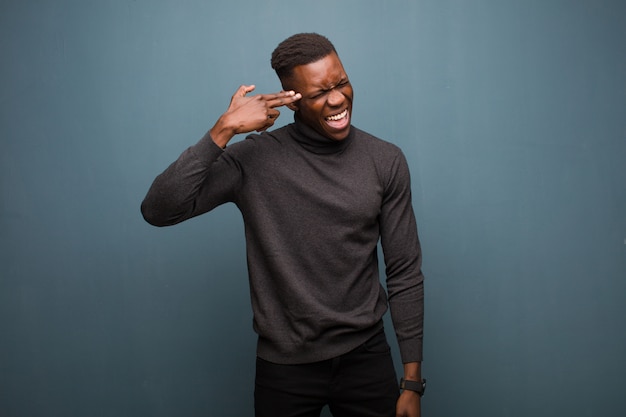 Young man looking unhappy and stressed, suicide gesture making gun sign with hand, pointing to head against grunge wall