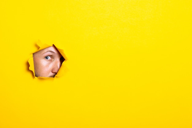 Young man looking with an eye into the hole of torn yellow paper