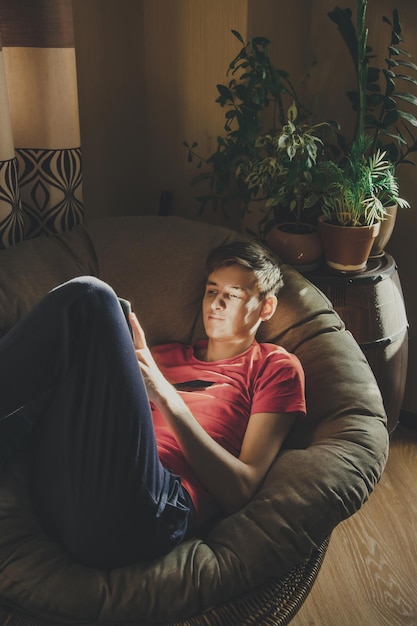 若い男は彼の顔に太陽のまぶしさで自宅の椅子に横たわっている彼のスマートフォンを見ます