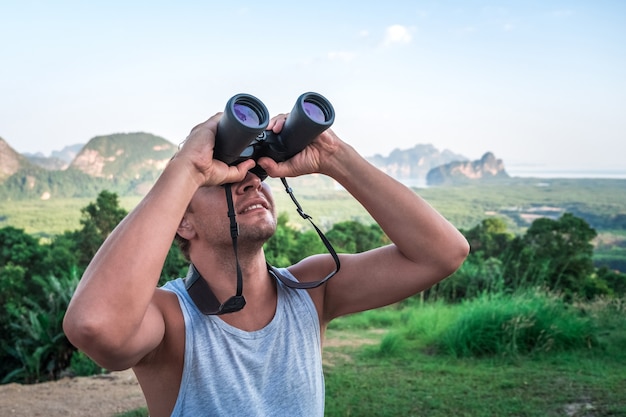 A young man looks into the sky through binoculars. A traveler on the background of wild nature.