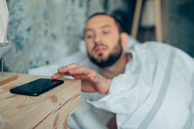 Young man lying in bed in the morning and switching off the alarm clock in his phone