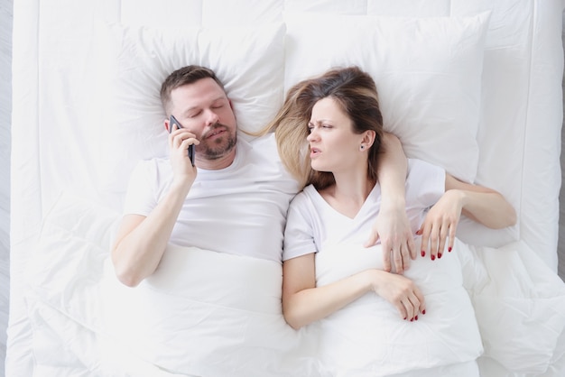 Young man lying in bed with his wife and talking on cell phone top view suspicion of treason