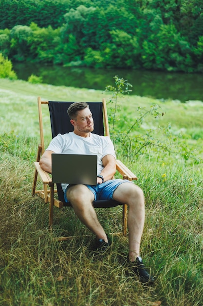 Young man lying on a chair in nature and working with a laptop resting alone thoughtfully looking up A man on a trip in the countryside the concept of people's lifestyle