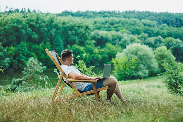 Молодой человек, лежащий на стуле на природе и работающий с ноутбуком, отдыхающий в одиночестве, вдумчиво глядя на человека в поездке по сельской местности, концепцию образа жизни людей