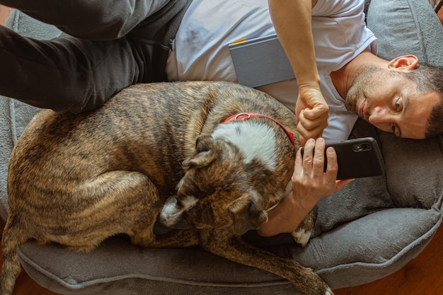 Молодой человек лежит рядом со своей собакой с книгой на груди и проверяет свой смартфон Малая глубина резкости