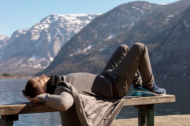 Giovane uomo sdraiato all'aperto su una panchina e godersi le montagne neve bel tempo cielo blu sole bellissimo paesaggio tempo con te stesso sognando relax salute mentale turismo vacanze viaggi