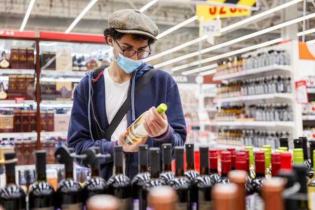 Молодой человек в медицинской маске, очках и кепке выбирает алкоголь в магазине. Депрессия и праздники во время пандемии коронавируса.