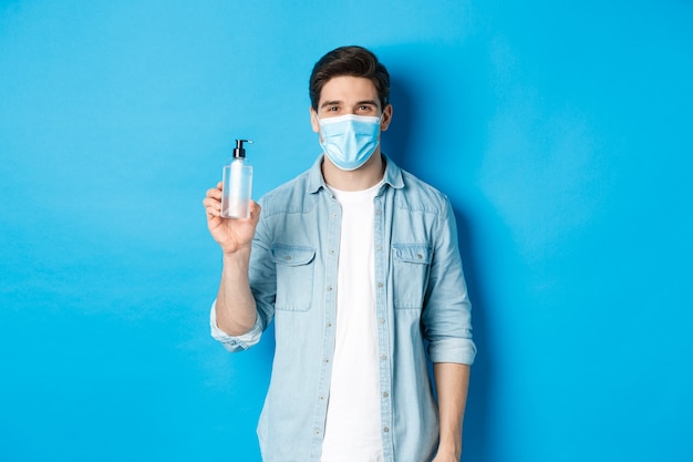 Young man in medical mask showing hand sanitizer, standing against blue wall