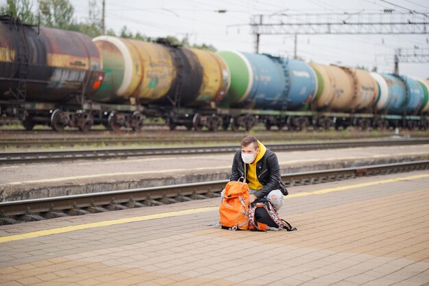 Young man in medical mask squats on platform waiting for train Male passenger in protective mask with backpacks sitting on railroad platform in waiting for train ride during coronavirus pandemic