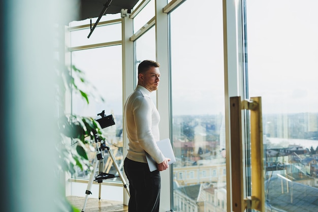 モダンなワークスペースの若い男性が大きな窓の近くに立っている ラップトップを手にしたカジュアルな服装の男性 リモートワーク 素敵な若いマネージャー