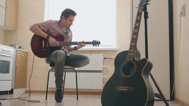 Young man musician composes music on the guitar and play in the kitchen, other musical instrument in the foreground, sitting