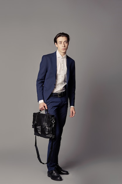 Young man in navy suit with black business bag over gray wall