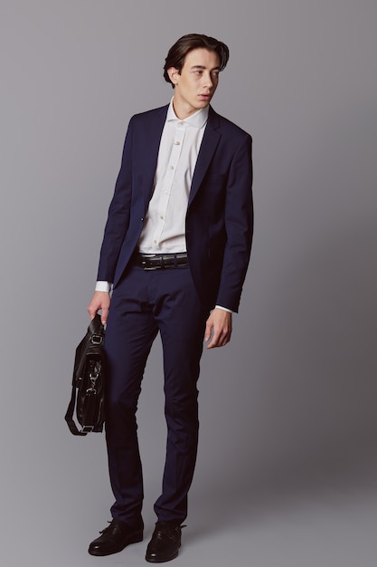 Young man in navy suit with black business bag over gray wall