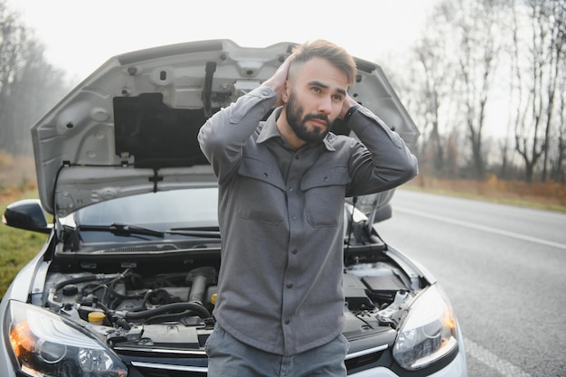 A young man near a broken car with an open hood on the roadside