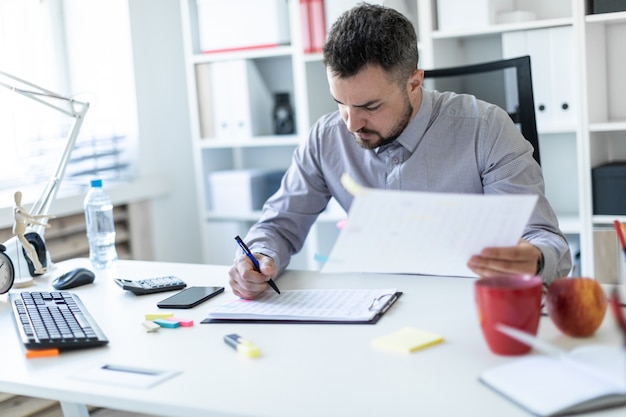 A young man in the office sits at a table, holds a pen in his hand and works with documents.