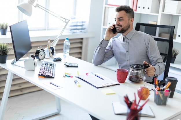 A young man in the office sits at a table, talking on the phone and holding a coffee pot.