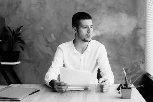 Young man in the office smokes a cigarette and exhales smoke while looking out the window thinking about the problem business crisis and businessman