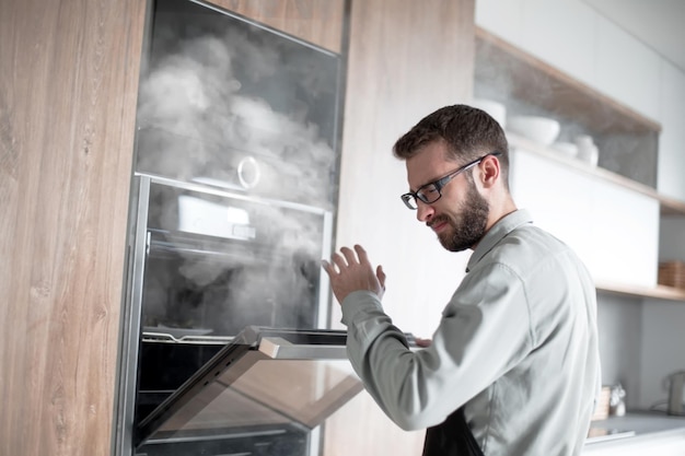 Young man opening electric oven in his kitchen