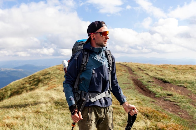 オレンジ色のジャケットとメガネの若い男がトレッキングスティックで山を歩いています