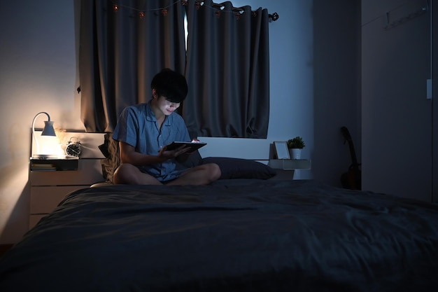 Young man in pajamas sitting in his comfortable bed and using digital tablet