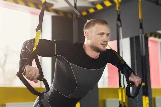 Young man performing TRX training in gym. Muscular guy doing exercise for biceps with elastic rope, copy space