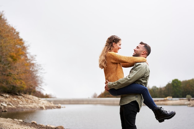 Young man picking up his girlfriend by the lake in fall