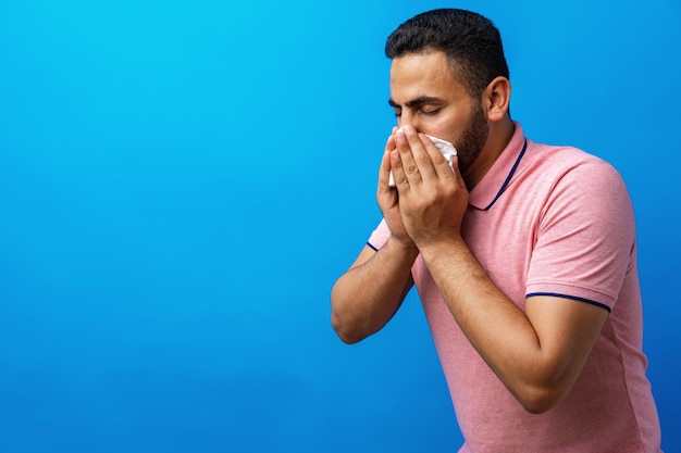 Young man in pink shirt with allergy or cold blowing his nose in a tissue against blue background
