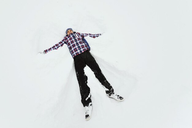 A young man in a plaid outerwear or jacket lies on the snow and makes the figure of an angel. Winter fun