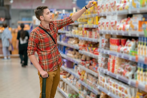 格子縞のシャツを着た若い男性がスーパーマーケットで食料品を買います。棚から商品を取り出す男性