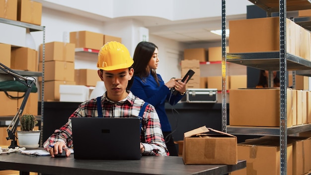 Young man planning order shipment in storehouse, using laptop to do stock logistics. Storage room employee working with products and merchandise, supply chain distribution packs.