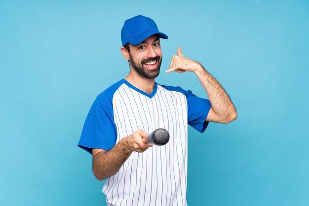 Young man playing baseball over isolated blue making phone gesture and pointing front