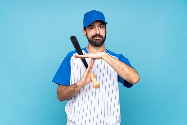 Young man playing baseball over isolated blue making time out gesture