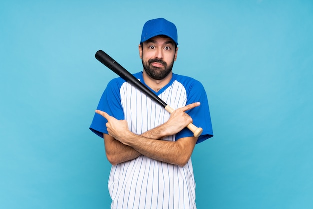 Young man playing baseball over isolated blue  pointing to the laterals having doubts