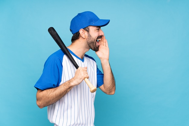 Young man playing baseball over isolated blue  shouting with mouth wide open to the lateral