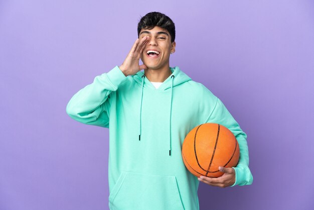 Young man playing basketball over isolated purple background shouting with mouth wide open