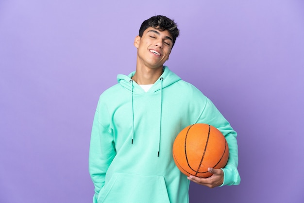 Молодой человек играет в баскетбол над изолированной фиолетовой стеной, смеясь
