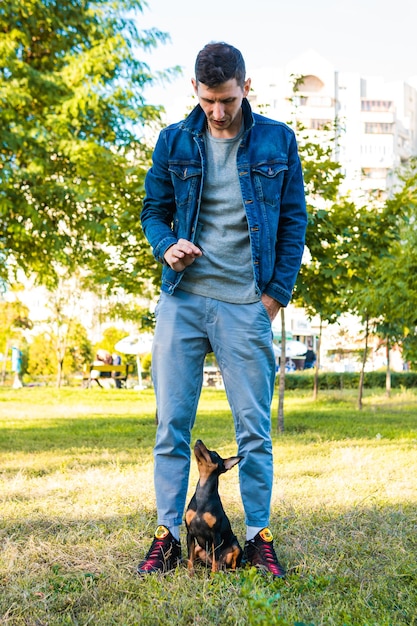 Young man playing and training his little dog outdoors. Black and Tan miniature pinscher female dog with owner in a park