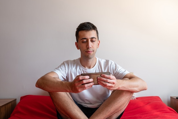 Young man playing with his smartphone sitting on the bed.