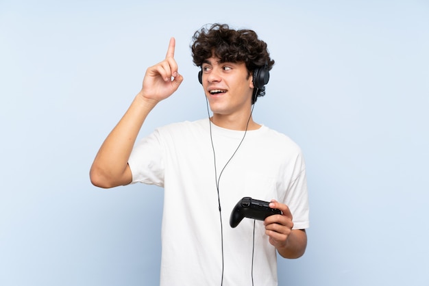 손가락을 들어 올리는 동안 솔루션을 실현하려는 격리 된 파란색 벽 위에 비디오 게임 컨트롤러를 가지고 노는 젊은 남자
