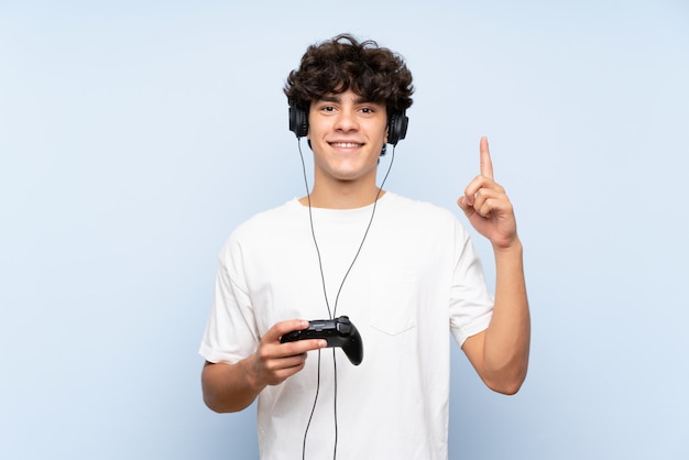 Young man playing with a video game controller over isolated blue wall pointing up a great idea