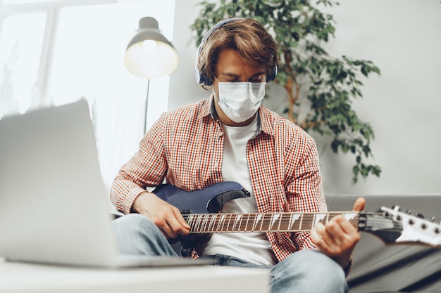 Young man plays guitar at home in medical mask