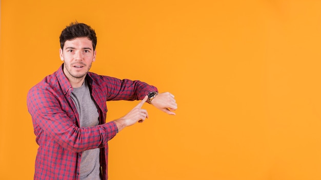 Young man pointing at wrist watch and looking at camera against orange background