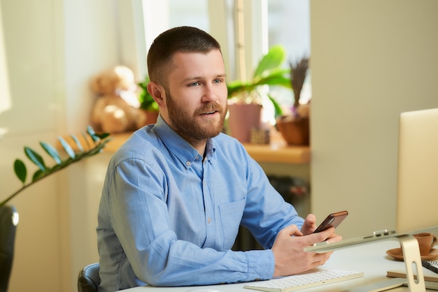 A young man poses sitting at a white desk in front of a laptop computer and holds a smartphone