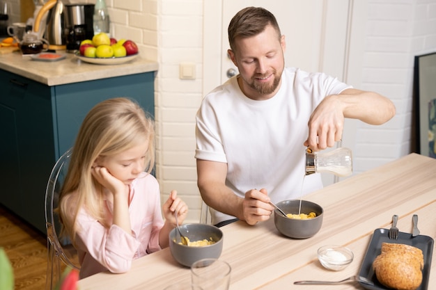 彼のかわいい小さな娘と一緒に朝食をとりながら、ミューズリーまたはコーンフレークと灰色の磁器のボウルに新鮮な牛乳を注ぐ若い男