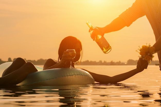 Young man pours wine into a glass to a woman who lies on inflatable ring in water during sunset