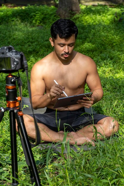 Young man practicing yoga asana balance meditating while standing on one leg on sports mat on green grass in park Using tablet for online class