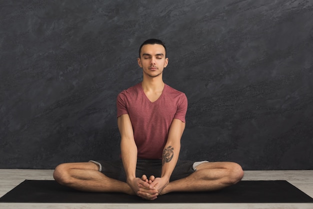 Young man practicing yoga, meditation exercises. Guy does lotus pose for relaxation, sitting with closed eyes on mat at gym. Healthy lifestyle, copy space