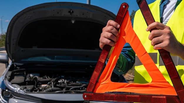 Young man preparing a red triangle to warn other road users car breakdown or engine failure stop at countryside street Male driver standing near a broken car with open up hood